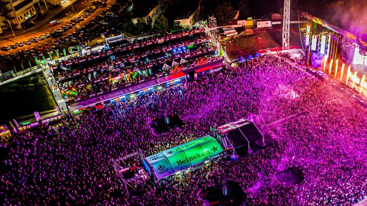 A large crowd of people is in front of a stage with pyrotechnics where a DJ is performing. The Heineken sponsor logo is in the middle.