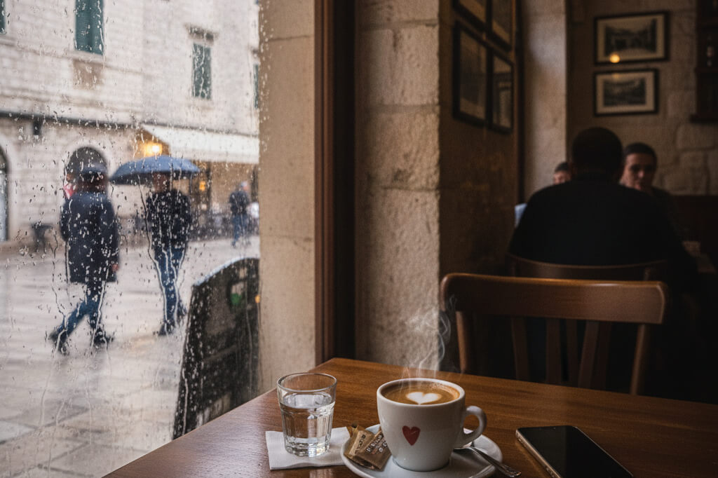 A cozy café interior with a steaming cup of coffee on a wooden table, seen next to a rain-covered window overlooking people walking with umbrellas outside.